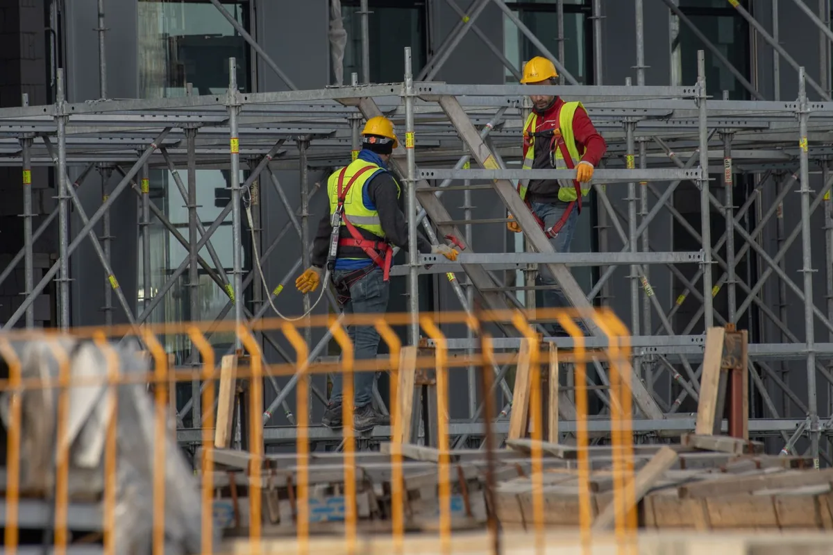 Two male general contractors in yellow hard hats and high visibility vests working together on scaffolding at an outdoor building site