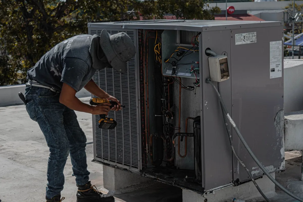HVAC technician in a bucket hat repairing an outdoor air conditioning unit on a rooftop, checking the system during a service visit