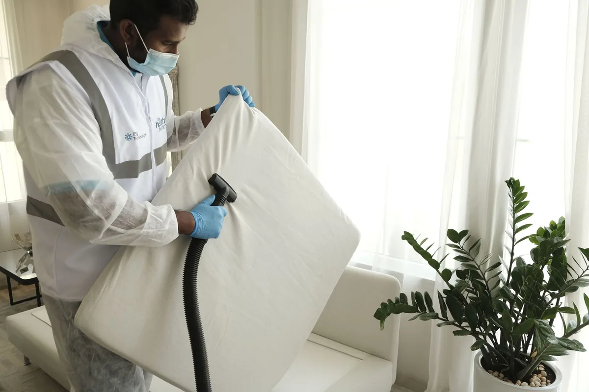 Male professional cleaner in protective uniform and blue gloves vacuuming a mattress during a deep cleaning service visit at a client home